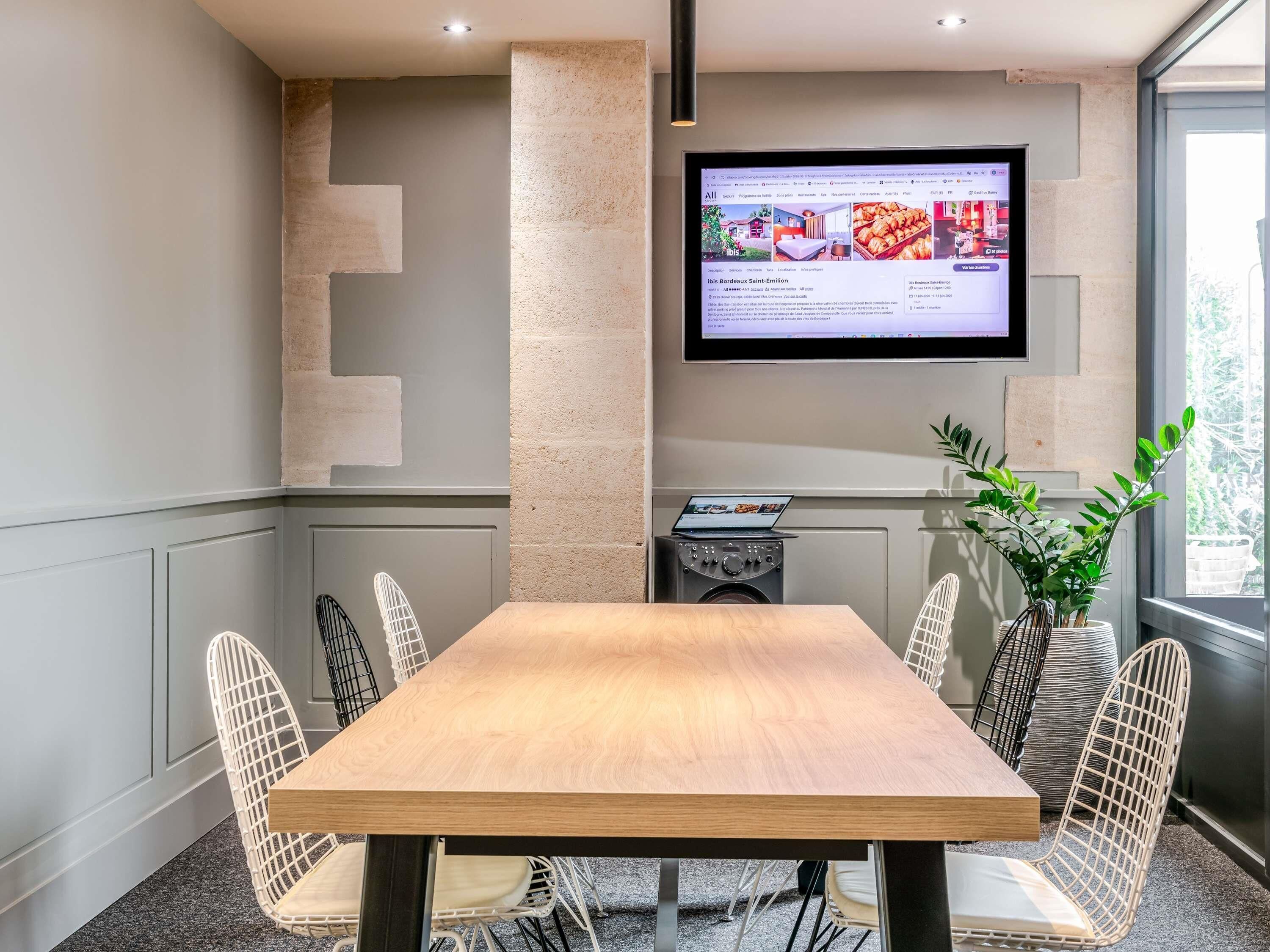 A conference room with a wooden table, white wireframe chairs, a mounted TV, and a potted plant.