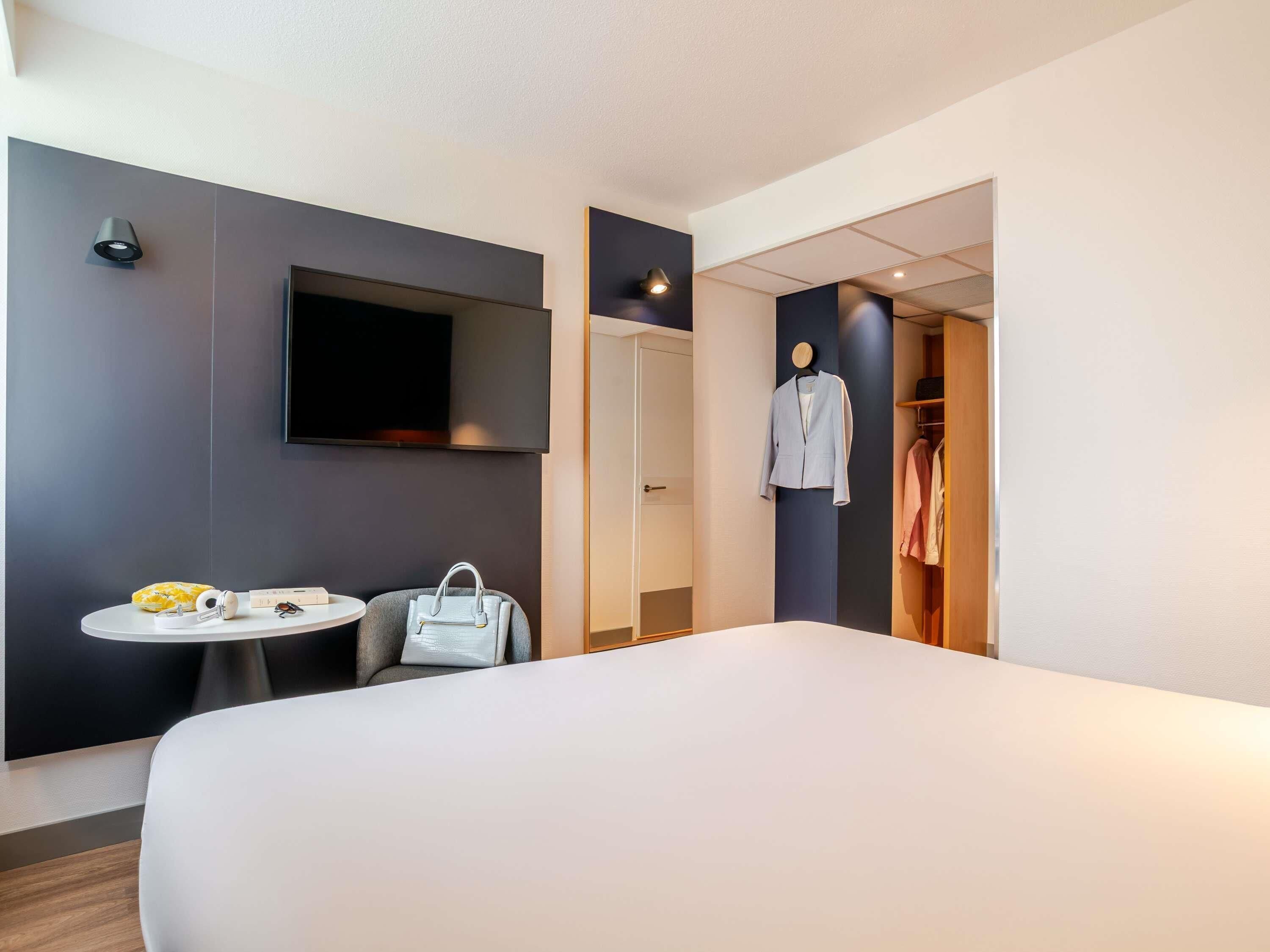 Quarto de hotel com uma cama, uma mesa redonda com uma cadeira, uma televisão fixada na parede, um cabideiro com um casaco e um guarda-roupa.