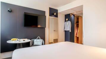 Quarto de hotel com uma cama, uma mesa redonda com uma cadeira, uma televisão fixada na parede, um cabideiro com um casaco e um guarda-roupa.