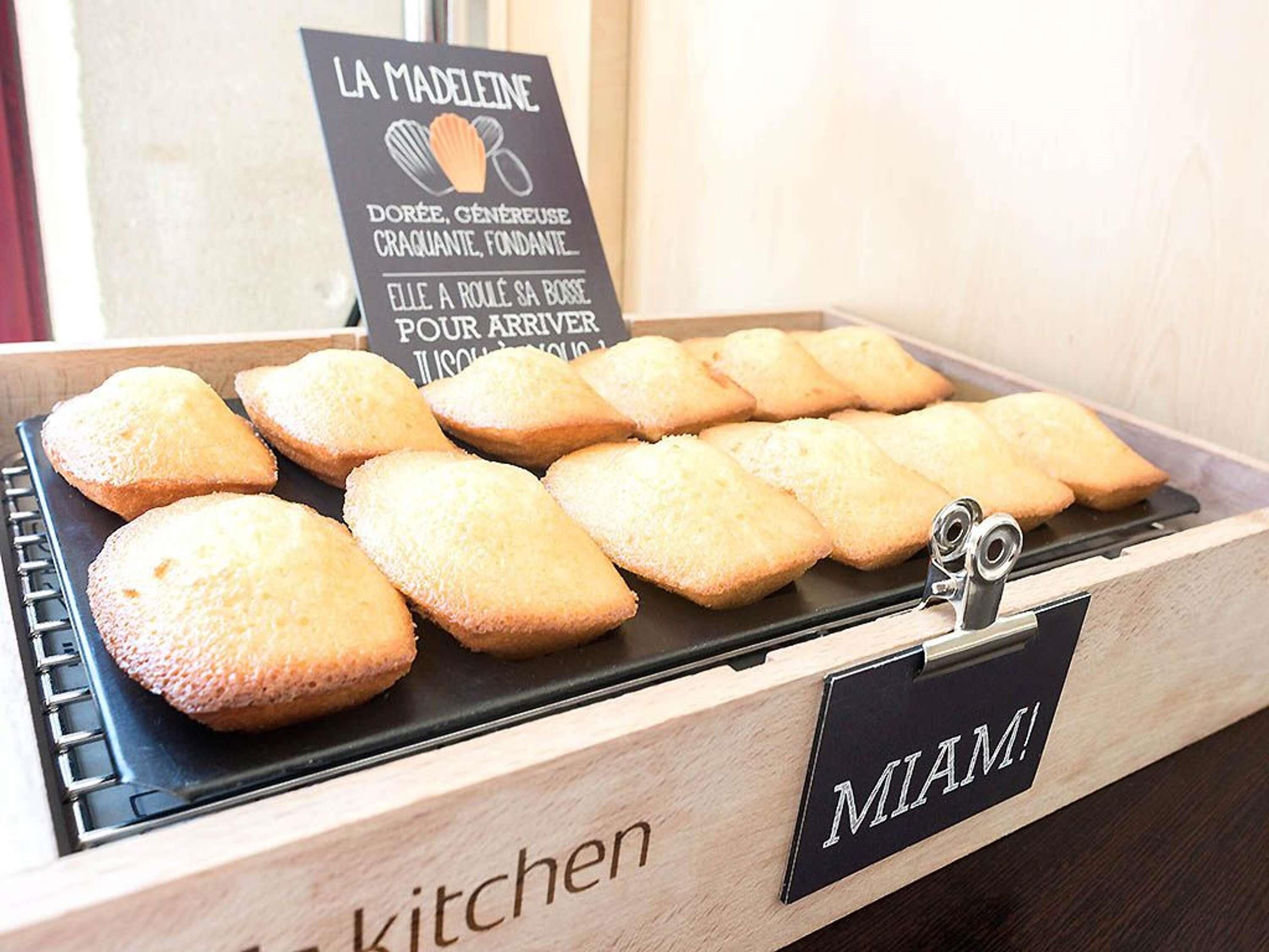 Uma bandeja de produtos de panificação numa caixa de madeira com a etiqueta 'MIAMI' e um letreiro acima que diz 'LA MADELEINE' e 'Dourada, generosa, crocante, macia'.