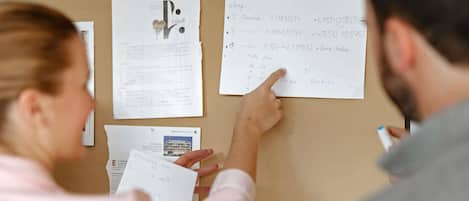 A person pointing at a bulletin board with papers pinned on it, including one with architectural plans and another with handwritten notes.