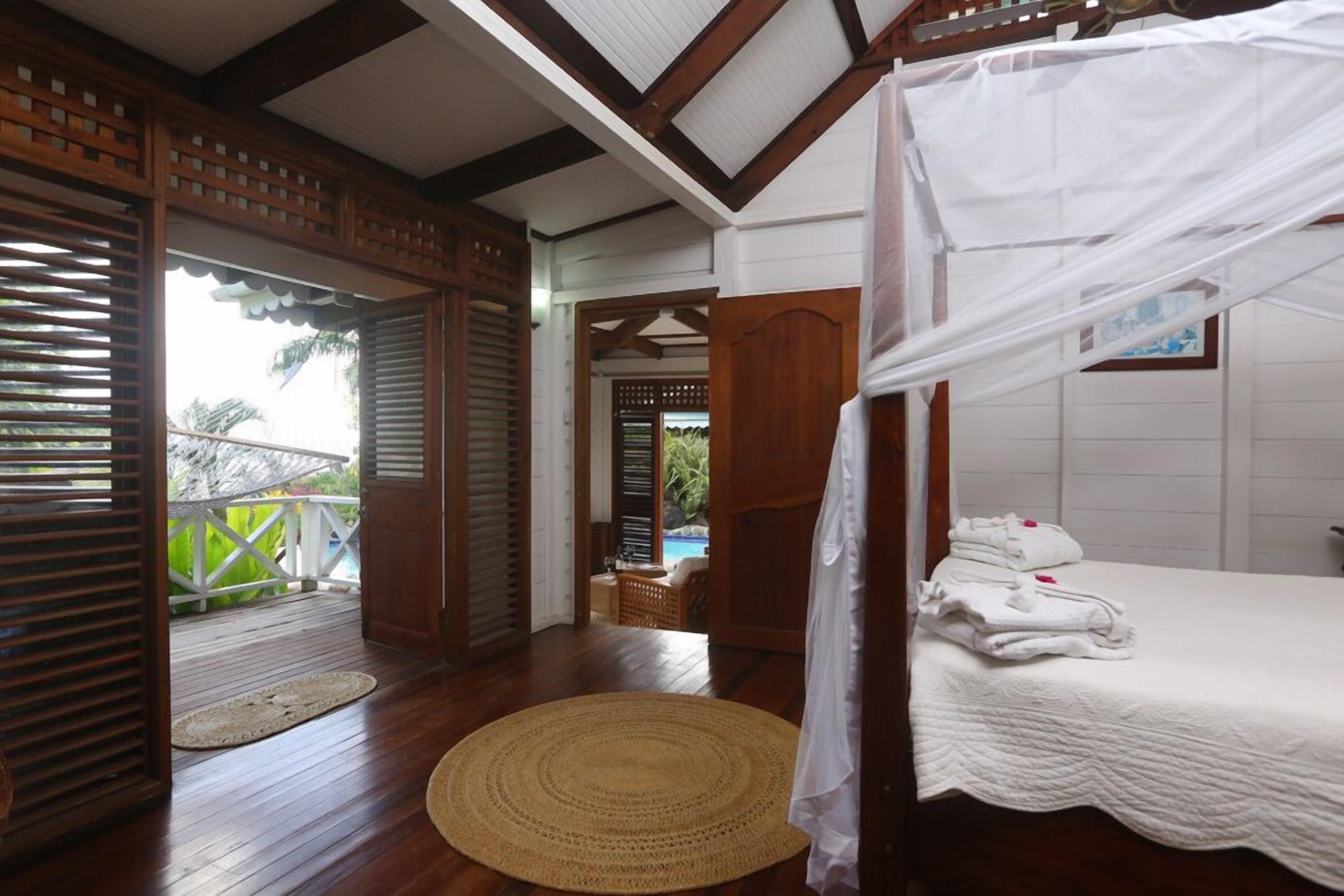 A bedroom with a canopy bed, wooden flooring, a woven mat, and a view of a pool and greenery.