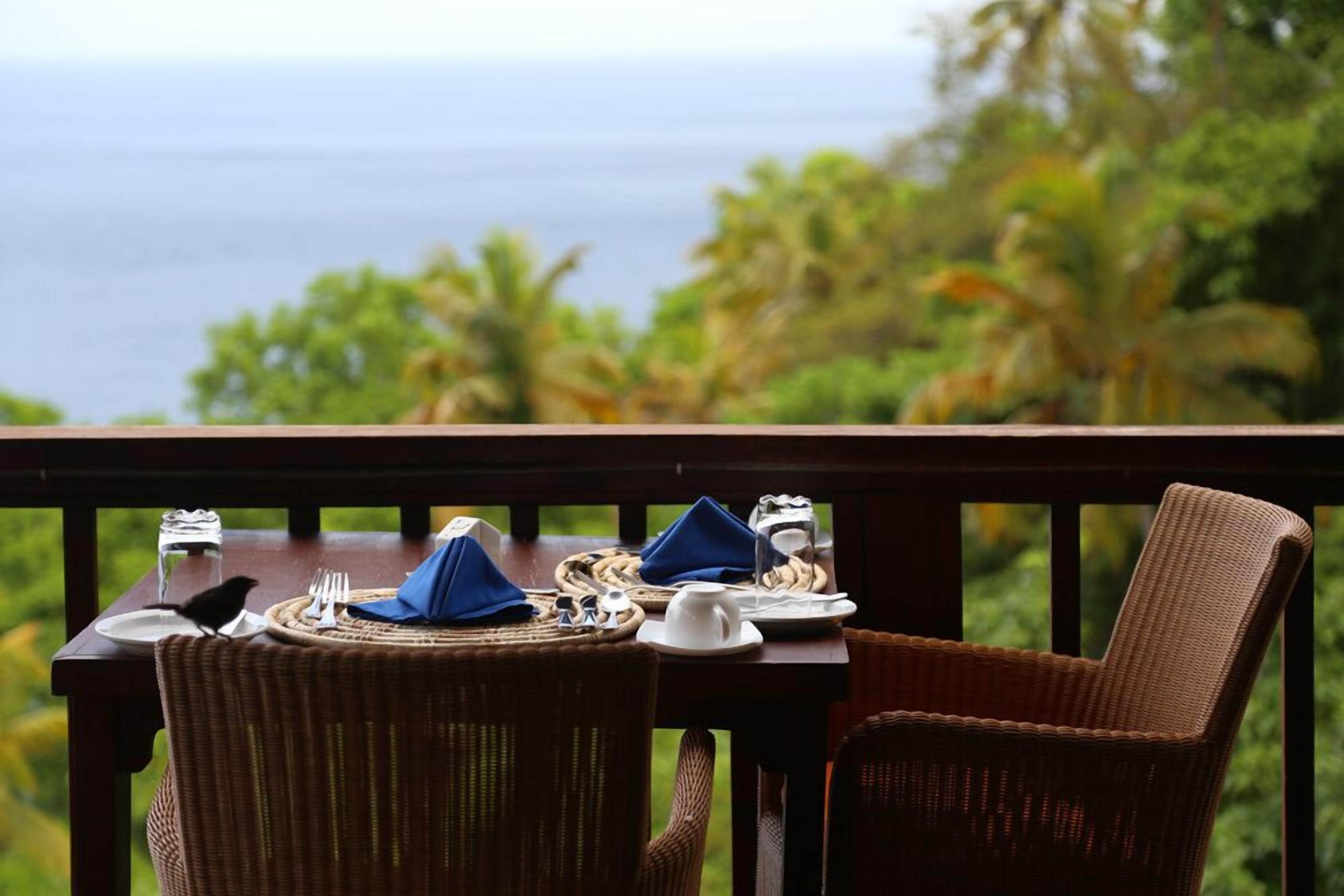 A dining table set with plates, cutlery, and a napkin on a balcony overlooking a scenic view of the ocean and palm trees.