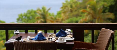 A dining table set with plates, cutlery, and a napkin on a balcony overlooking a scenic view of the ocean and palm trees.