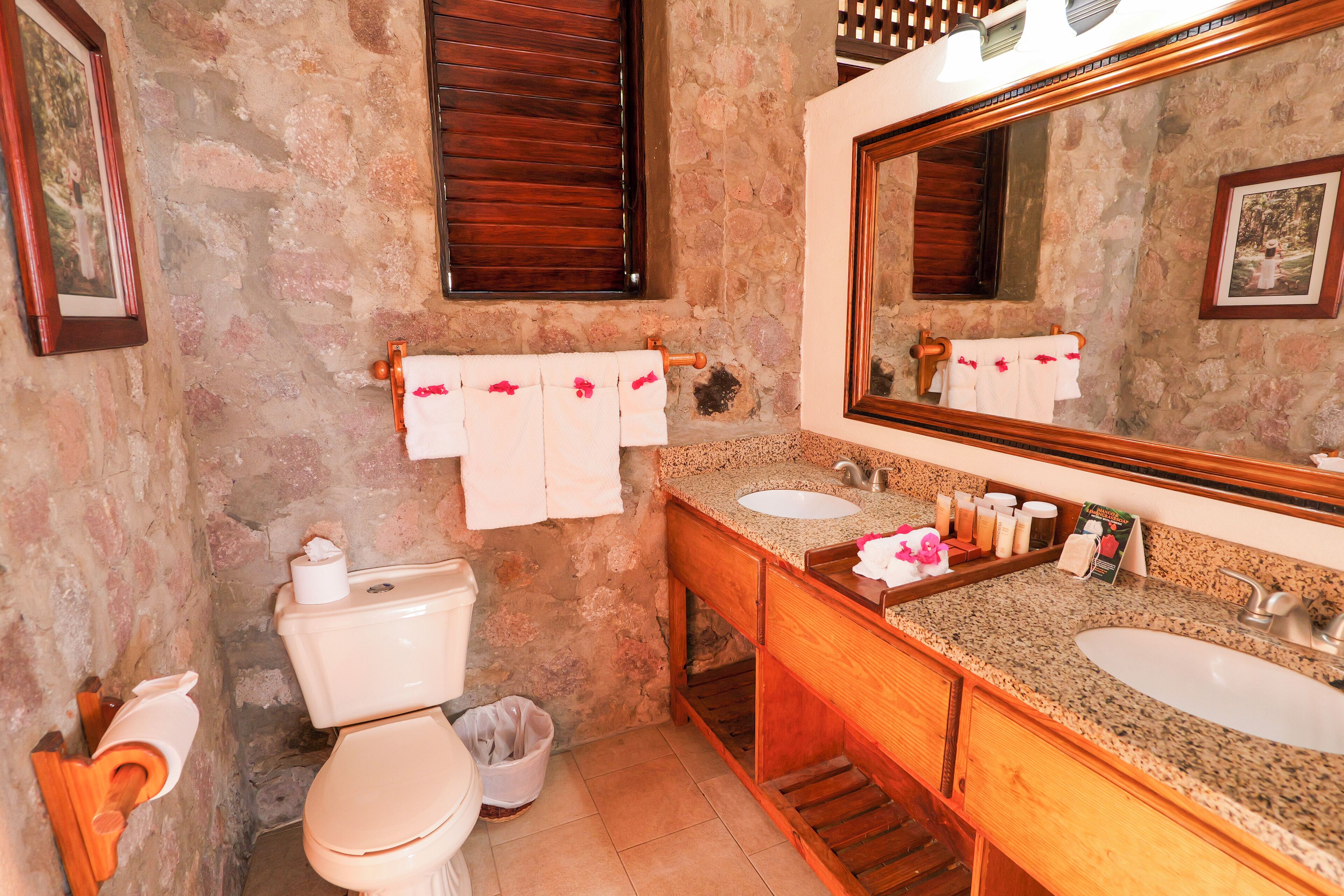 A bathroom with a stone wall, wooden vanity, two sinks, a toilet, and a large mirror.
