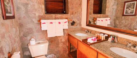 A bathroom with a stone wall, wooden vanity, two sinks, a toilet, and a large mirror.