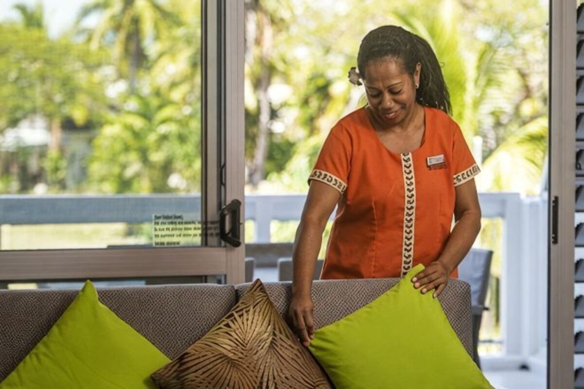 A person arranging pillows on a sofa in a room with a view of palm trees.