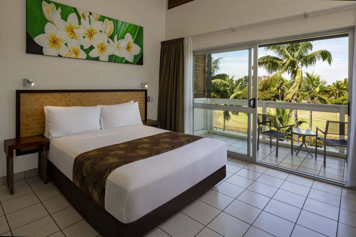 A hotel room with a large bed, a nightstand, a painting of flowers, and a balcony with a view of palm trees.