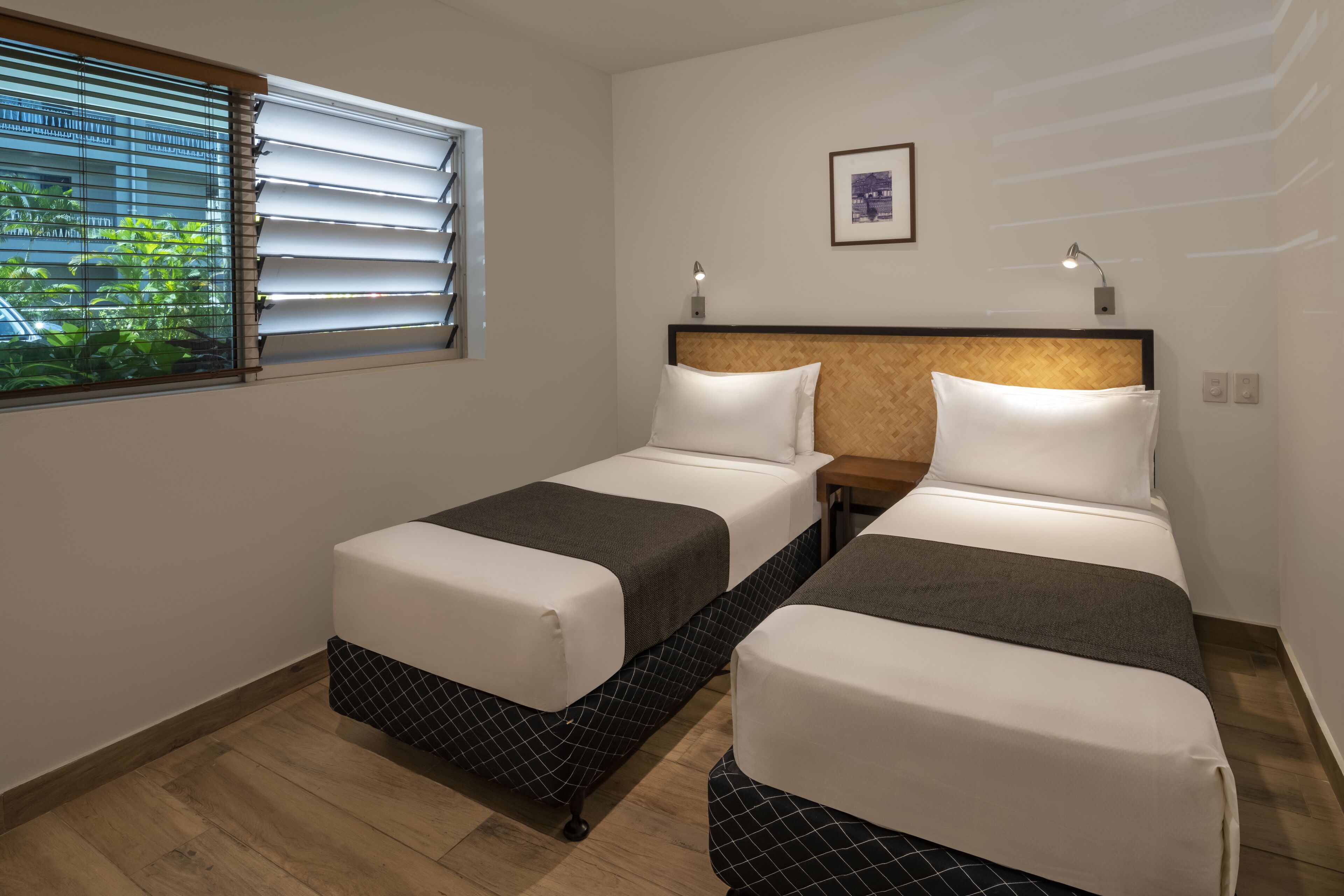 Una camera d'albergo con due letti, una finestra con persiane, un quadro appeso al muro e un comodino con una lampada.