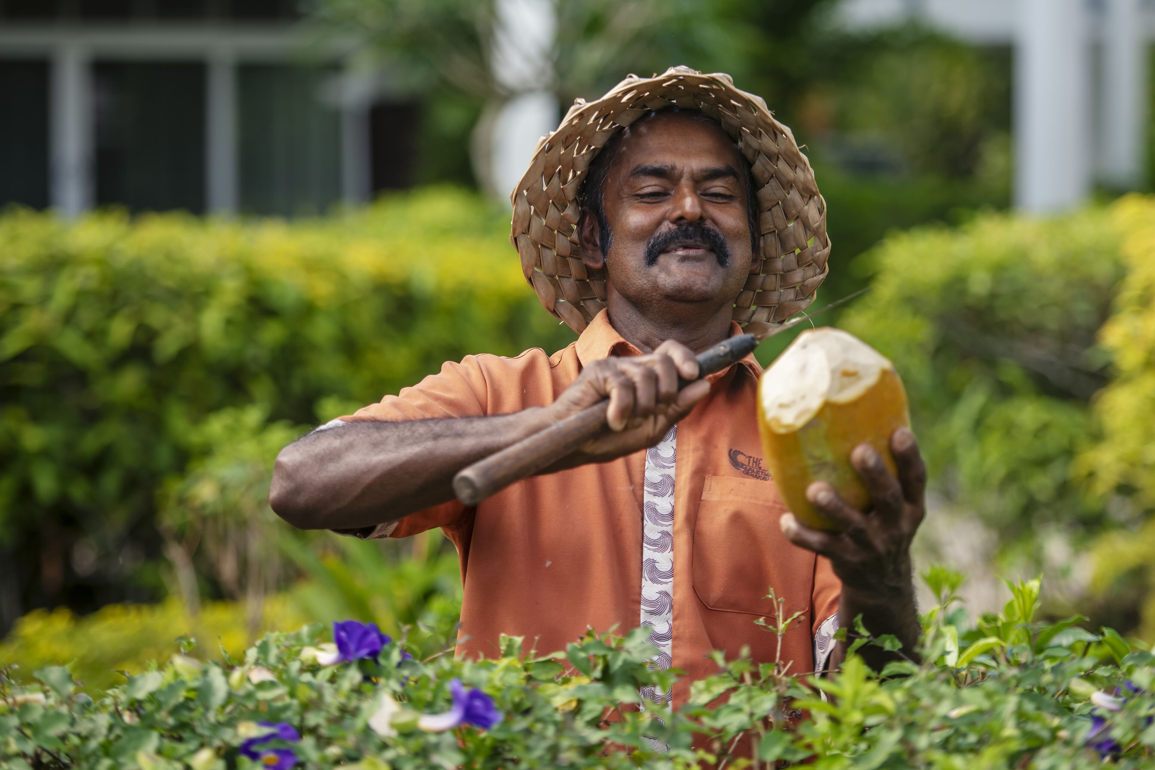 Una persona con una camisa naranja y un sombrero tejido sostiene un objeto amarillo grande, probablemente una fruta, en un jardín con follaje verde y un edificio al fondo.
