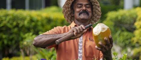 Una persona con una camisa naranja y un sombrero tejido sostiene un objeto amarillo grande, probablemente una fruta, en un jardín con follaje verde y un edificio al fondo.