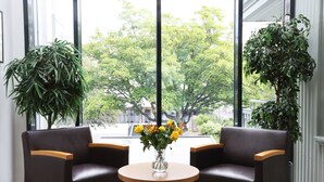 Una habitación con dos sillones de cuero, una pequeña mesa redonda y un florero con flores amarillas. Se ven ventanas grandes con vistas a árboles y edificios.