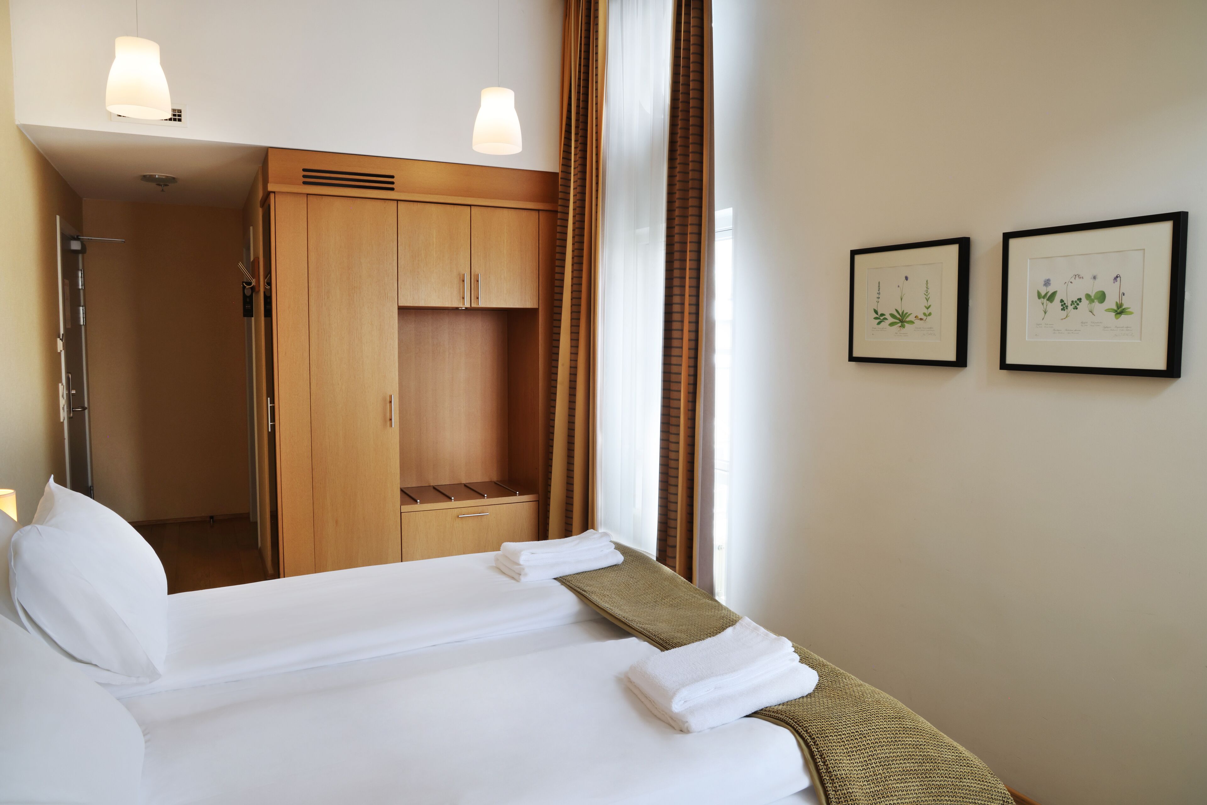 A hotel room with a bed, a wardrobe, a shower, and two framed botanical prints on the wall.
