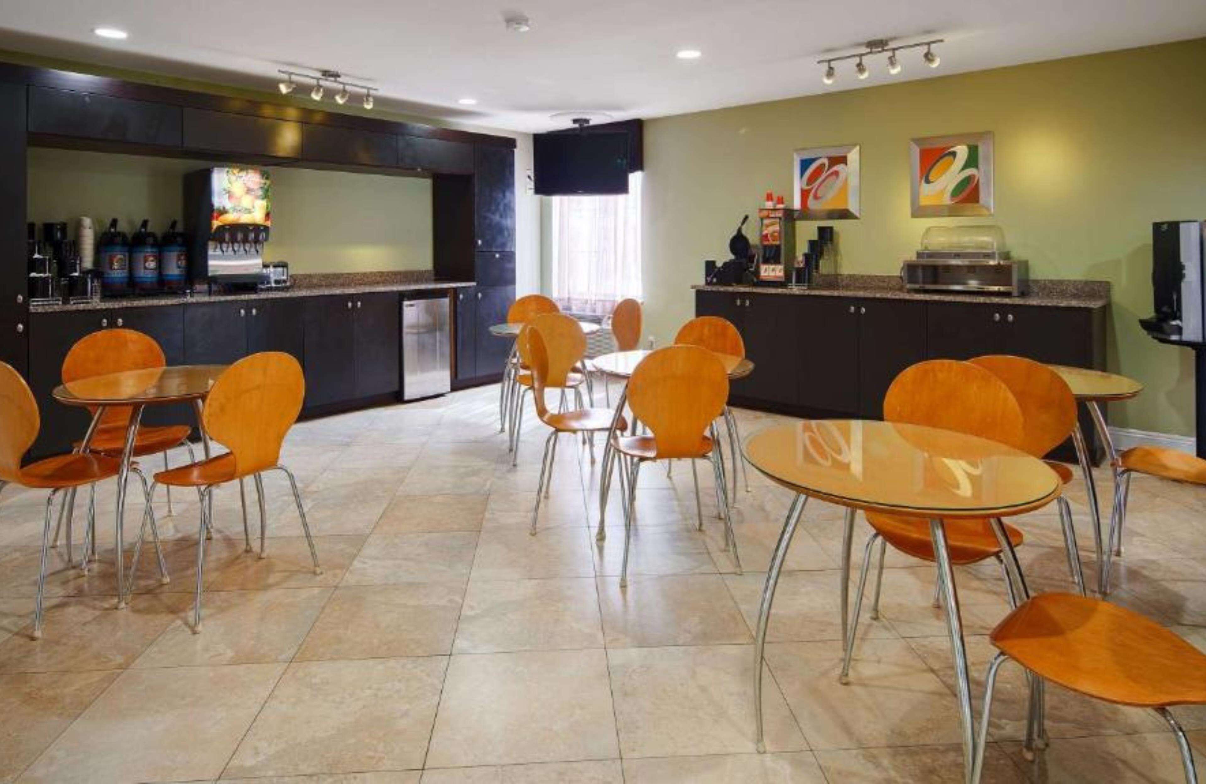 A hotel dining area with multiple tables and chairs, a kitchen area with a refrigerator and coffee machine, and artwork on the walls.