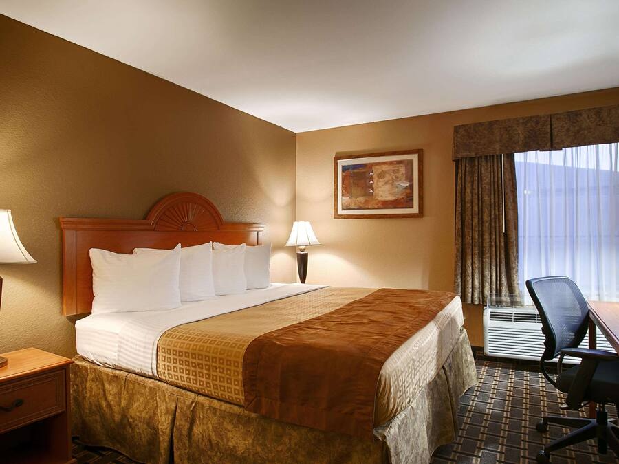 A hotel room with a large bed, two bedside lamps, a desk with a chair, and a window with curtains.