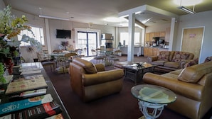 A spacious living area with leather sofas, a glass coffee table, and a dining table with chairs. There is a kitchen area in the background with cabinets and a flat-screen TV mounted above the counter.