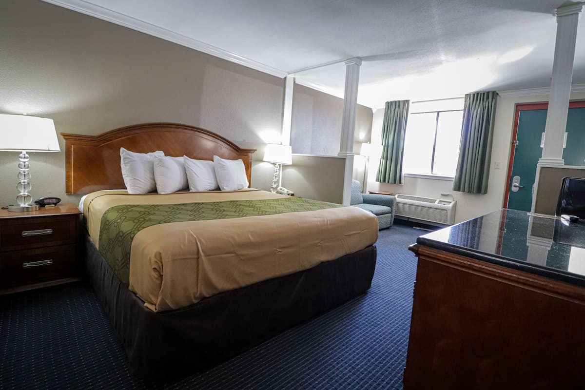 Habitación de hotel con una cama grande, una mesita de noche con lámpara, un escritorio con ordenador y una ventana con cortinas.
