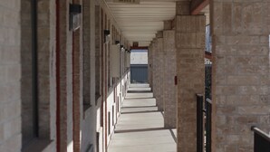 A covered corridor with a series of rooms on either side, a central exit sign, and a view of a building in the distance.