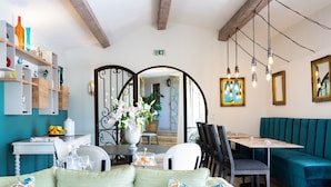 Un comedor con una mesa de madera, sillas y un banco de color verde azulado. Se observan estantes con objetos decorativos y un jarrón con flores a la izquierda.