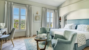 Un dormitorio con una cama, dos sillones, una mesita y vistas a la ciudad a través de amplios ventanales.