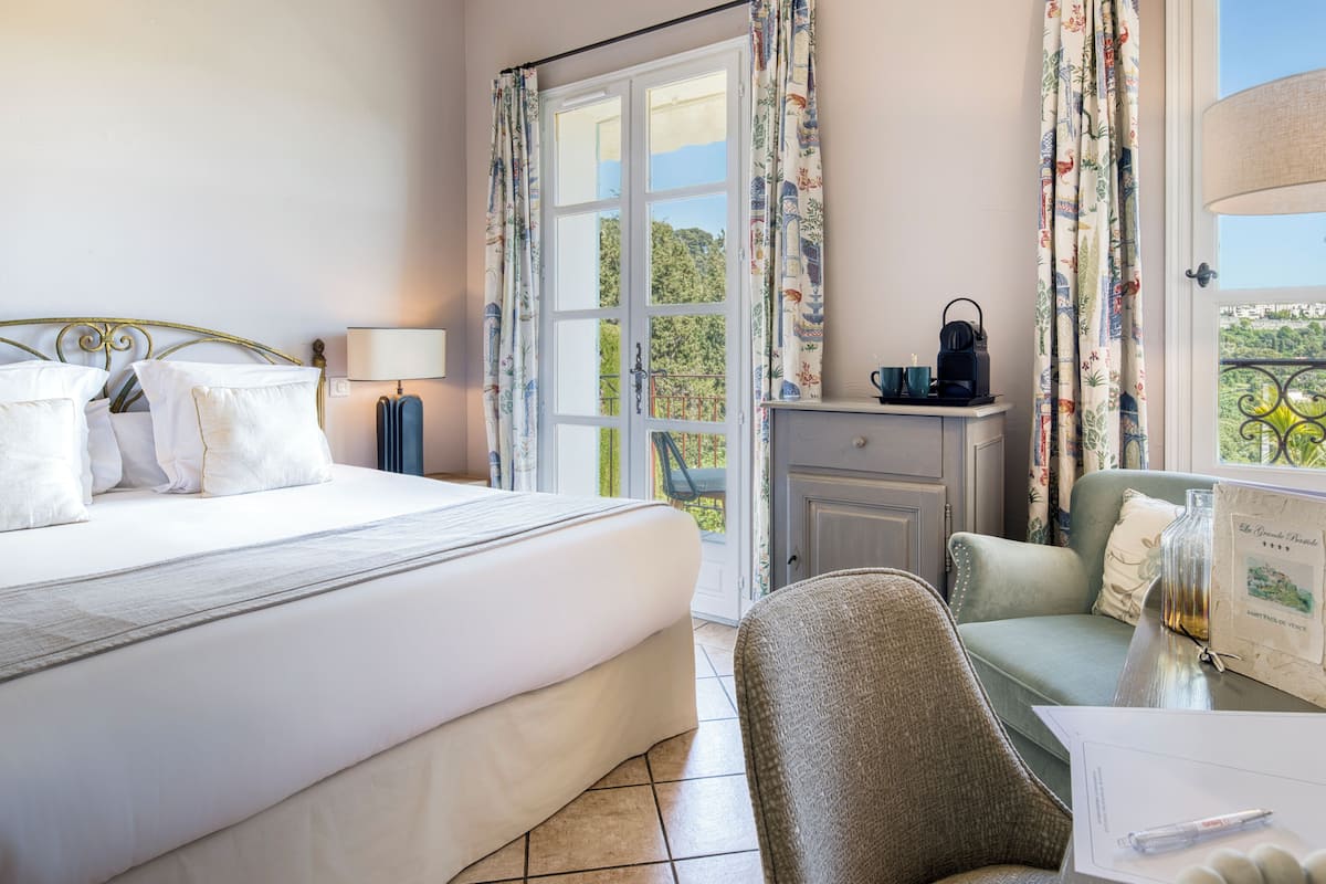 A hotel room with a bed, a desk with a lamp, a chair, a window with curtains, and a view of greenery.