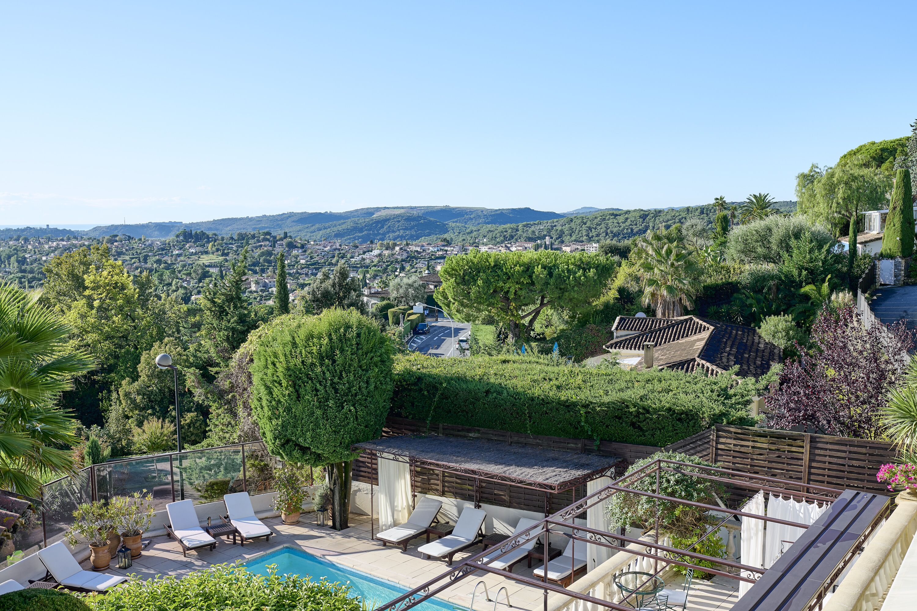 A rooftop pool area with lounge chairs, a pergola, and a view of a cityscape and hills.