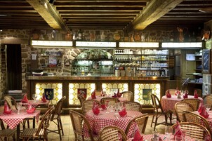 Un restaurante con sillas de mimbre y manteles a cuadros rojos y blancos, una zona de bar con varias botellas, y un fondo de pared de piedra.
