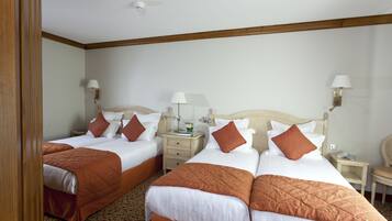 A hotel room with two single beds, each with a wooden headboard, white bedding, and orange throw pillows. There are bedside tables with lamps, a small nightstand, and a chair.