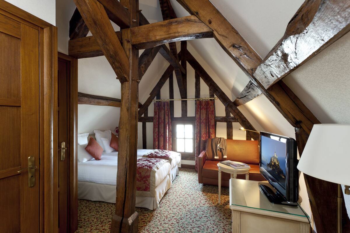 Een kamer met een hoge plafond, zichtbare houten balken, een bed met een bloemenlaken, een bank, een kleine tafel met een lamp en een televisie.