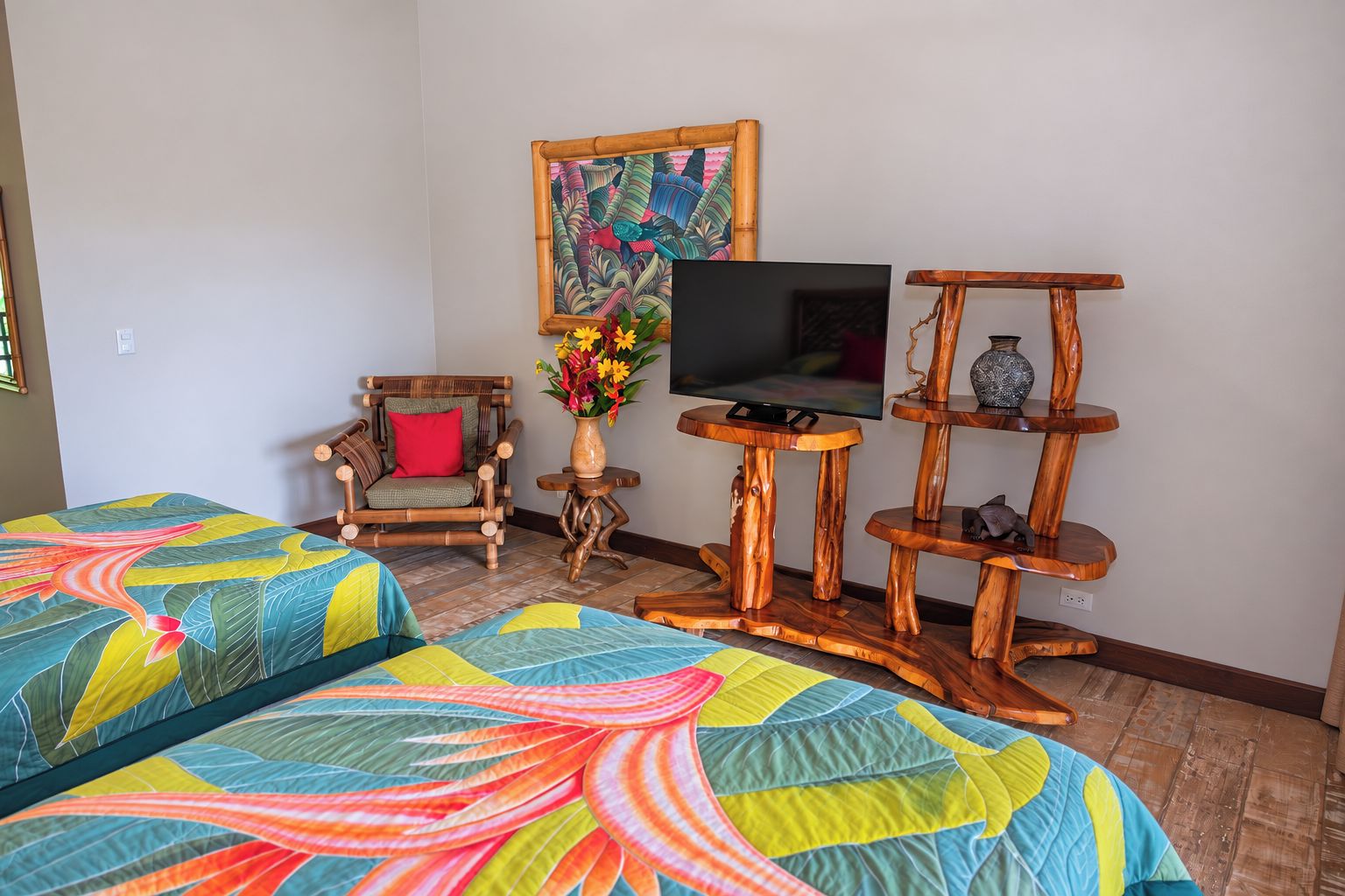 A room with two beds, a television on a wooden stand, a chair, and a decorative vase with flowers.