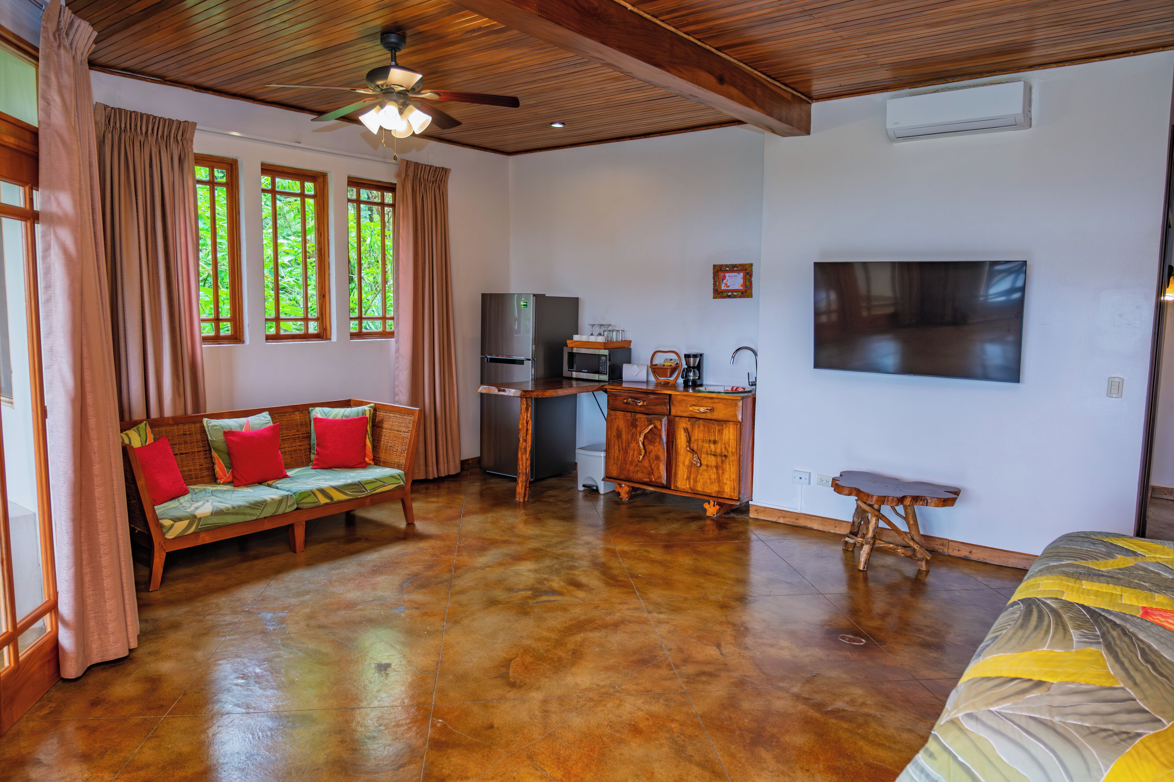 Amplia sala de estar con techo de madera, un sofá con cojines de colores, una mesa de comedor, una zona de cocina con refrigerador y microondas, y un televisor de pantalla plana grande montado en la pared.