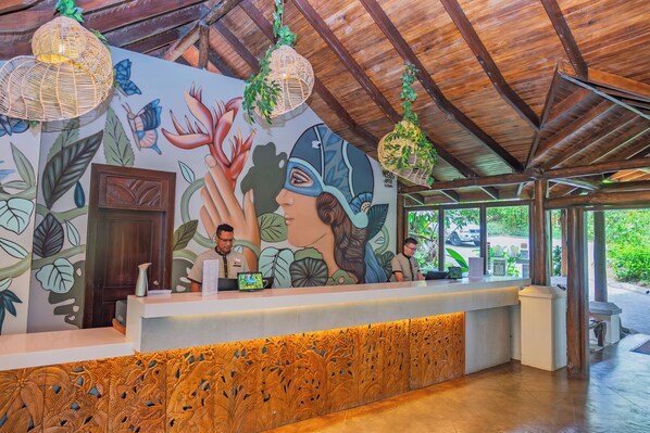 A reception area with a wooden ceiling, a mural with floral and bird designs, and a reception desk with two staff members. - Lost Iguana Resort and Spa (La Fortuna)