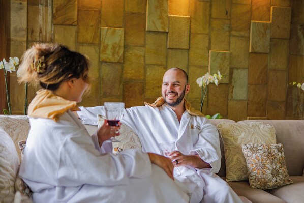 Un couple est assis sur un canapé beige agrémenté de coussins à motifs et tient des verres à vin.