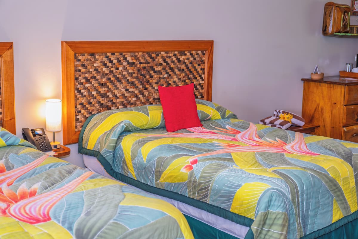 Une chambre avec un lit agrémenté d’une couverture aux couleurs vives, d’une tête de lit en bois, d’un oreiller rouge et d’une table de chevet avec une lampe et un téléphone.