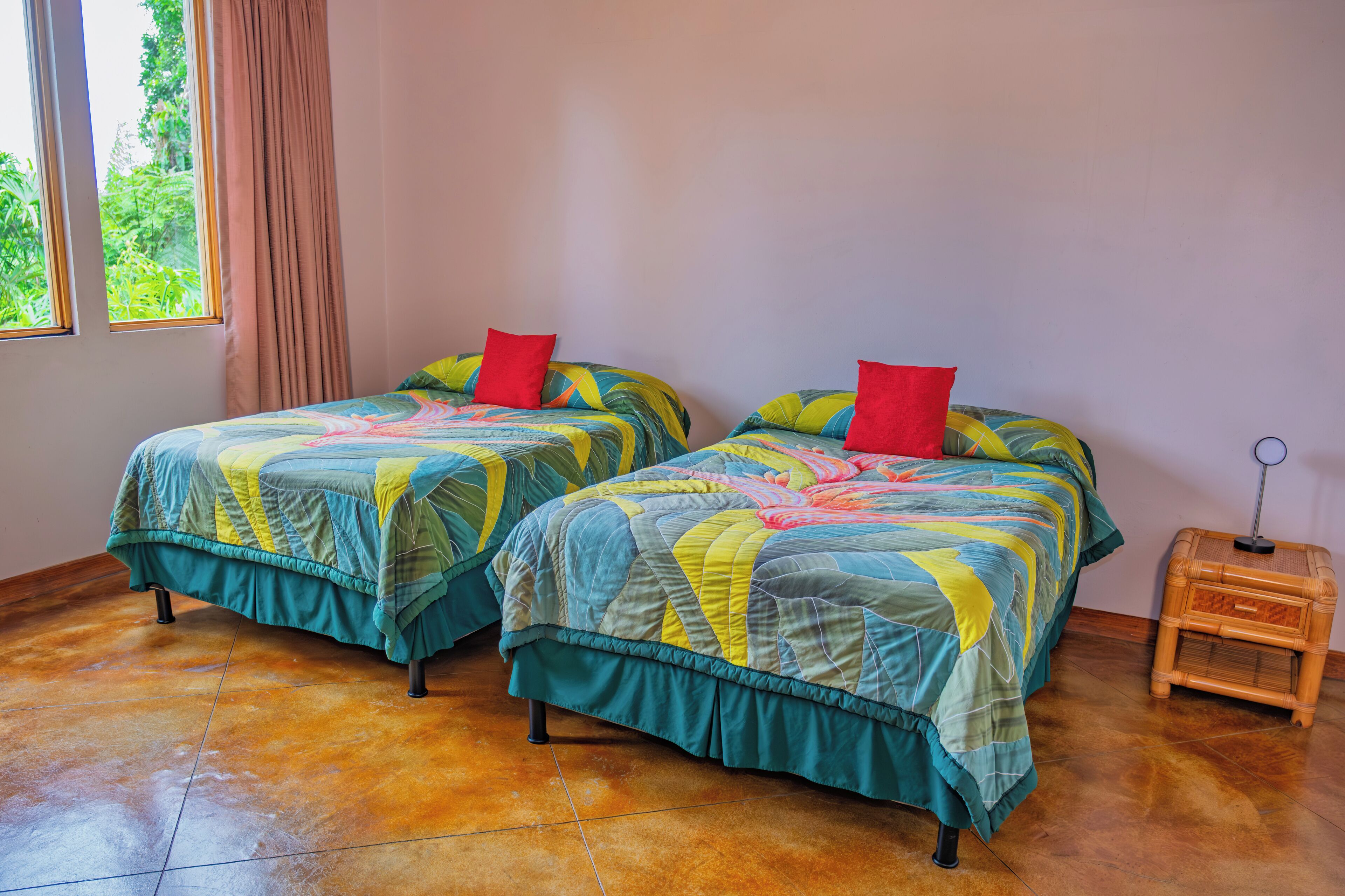 Two single beds with colorful quilts and red pillows, a small wooden nightstand, and a window with curtains.