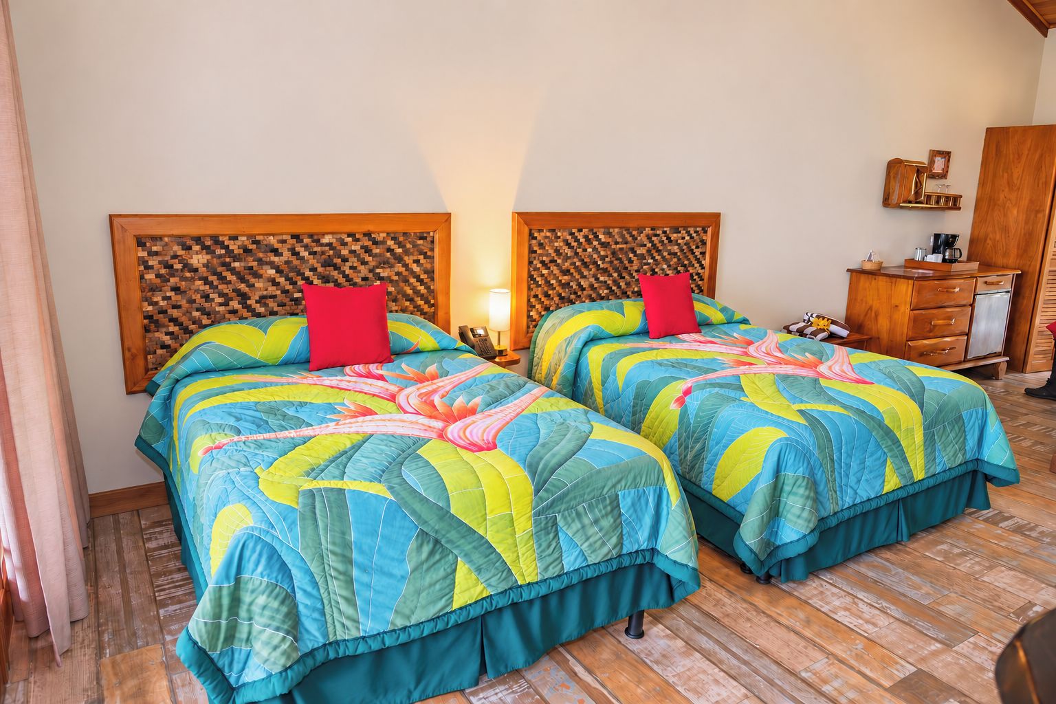 A room with two beds, each with a colorful quilt, a wooden headboard, a nightstand with a lamp, and a wooden dresser with a mirror.