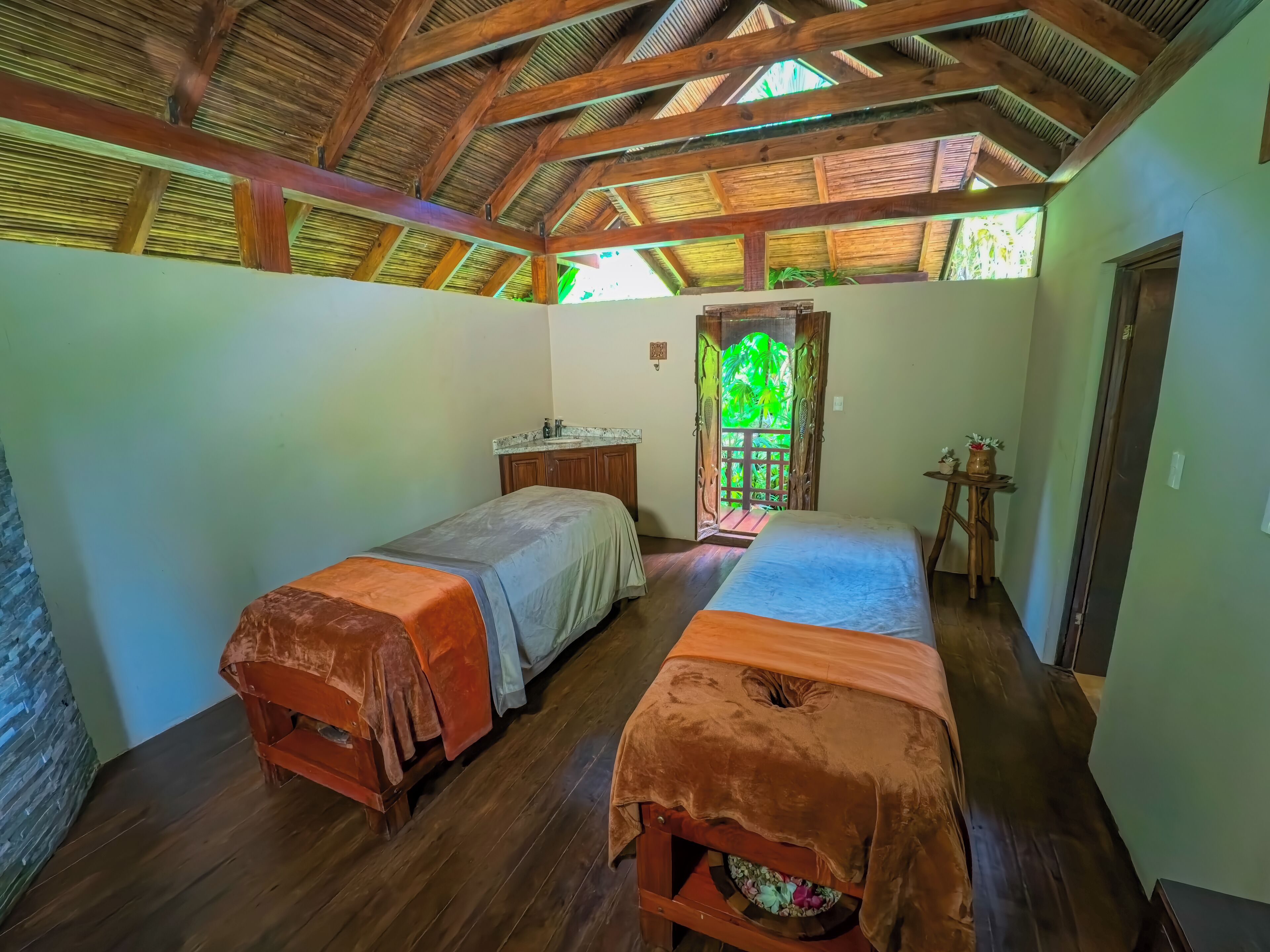 A room with two beds, a wooden ceiling, a small table, and a door leading to a balcony.