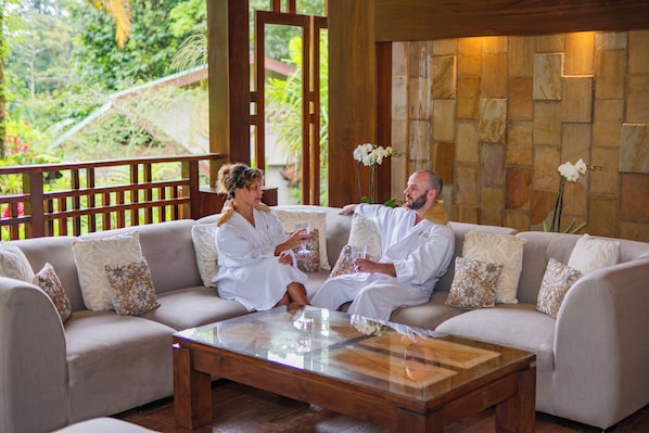 A couple in white robes sitting on a beige sofa with patterned cushions, in a room with a stone wall and a wooden railing overlooking greenery. - Lost Iguana Resort and Spa (La Fortuna)