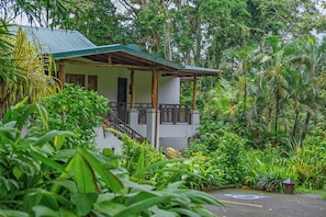 A two-story building with a green roof and wooden balcony railings, surrounded by lush tropical vegetation.
