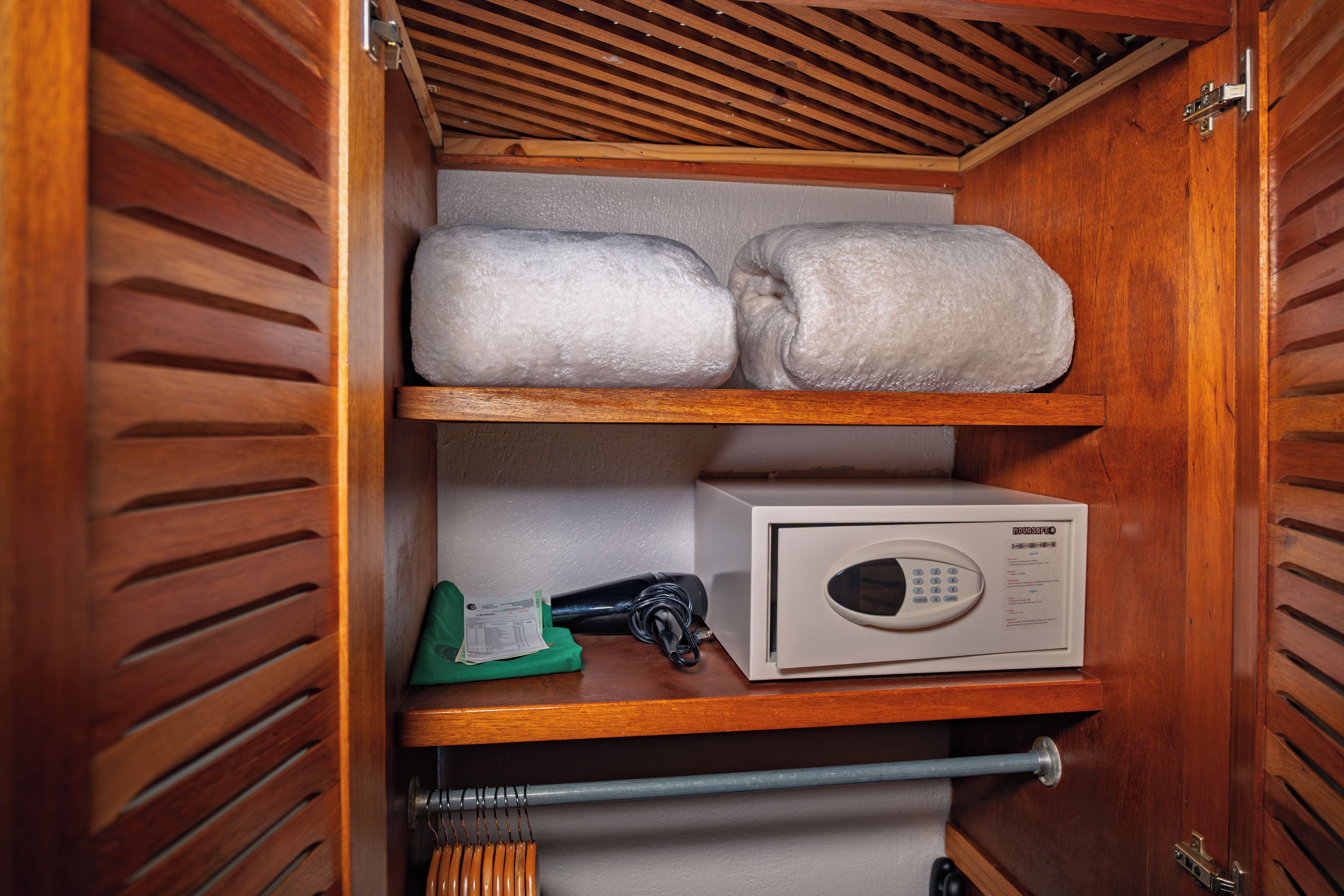 Un placard en bois avec deux étagères : sur la première, on aperçoit deux serviettes enroulées, et sur la seconde, un appareil blanc, qui pourrait être un four à micro-ondes, ainsi qu’un objet vert sur l’étagère inférieure.