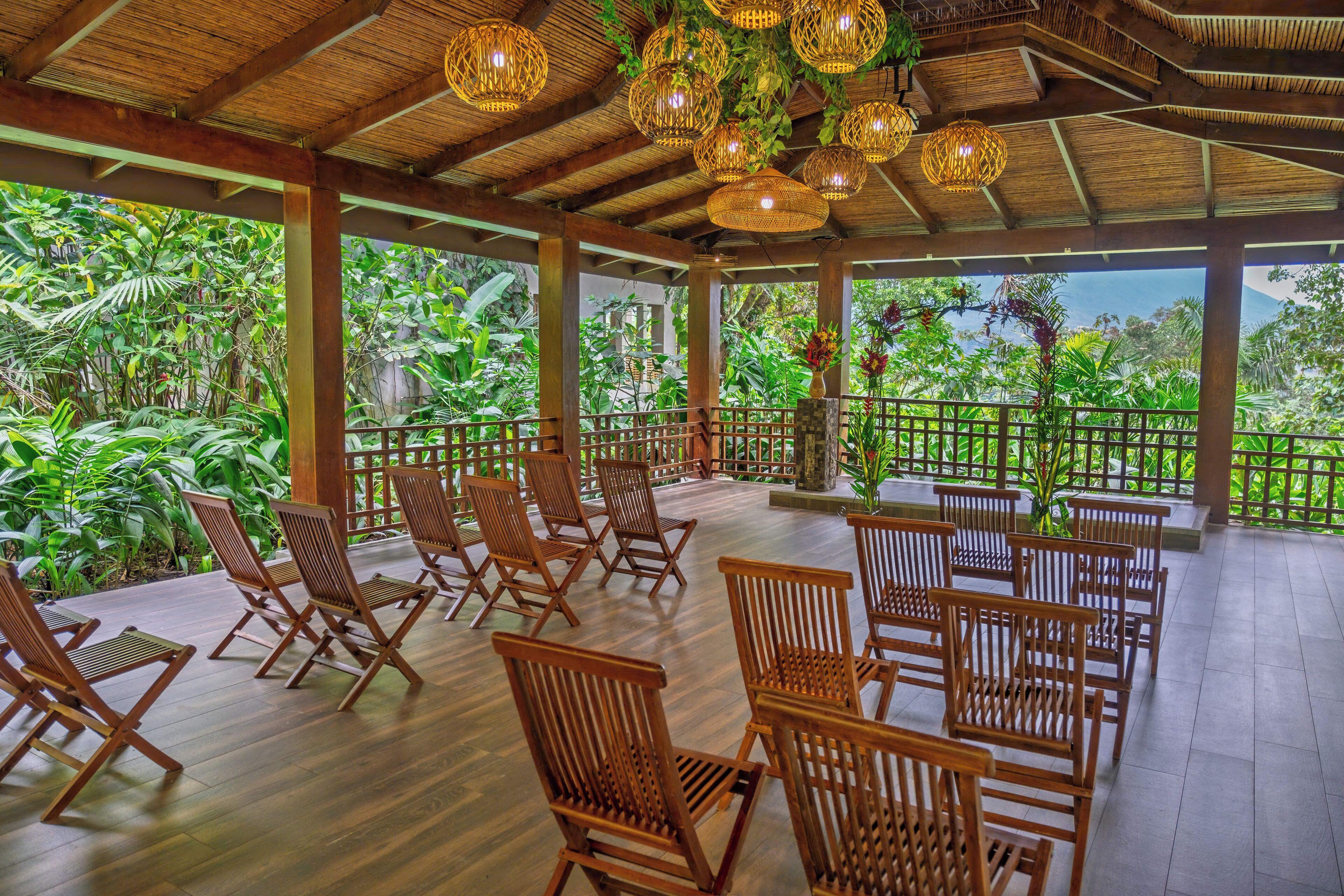 A covered outdoor seating area with wooden chairs and tables, surrounded by lush greenery and a view of the landscape.