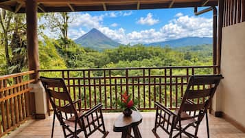 A wooden balcony with two rocking chairs and a small table, overlooking a lush landscape with a mountain in the distance.
