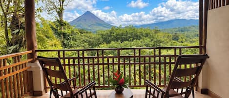 A wooden balcony with two rocking chairs and a small table, overlooking a lush landscape with a mountain in the distance.