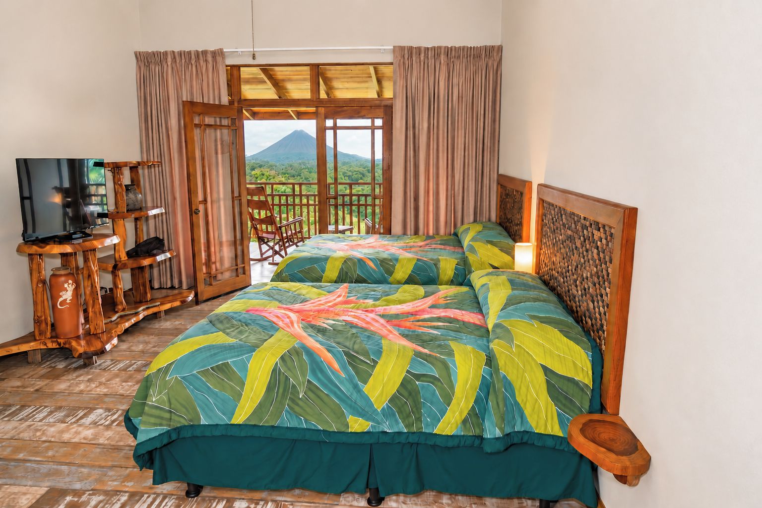 Habitación de hotel con cama, televisor, mesita de madera y balcón con vista a la montaña.