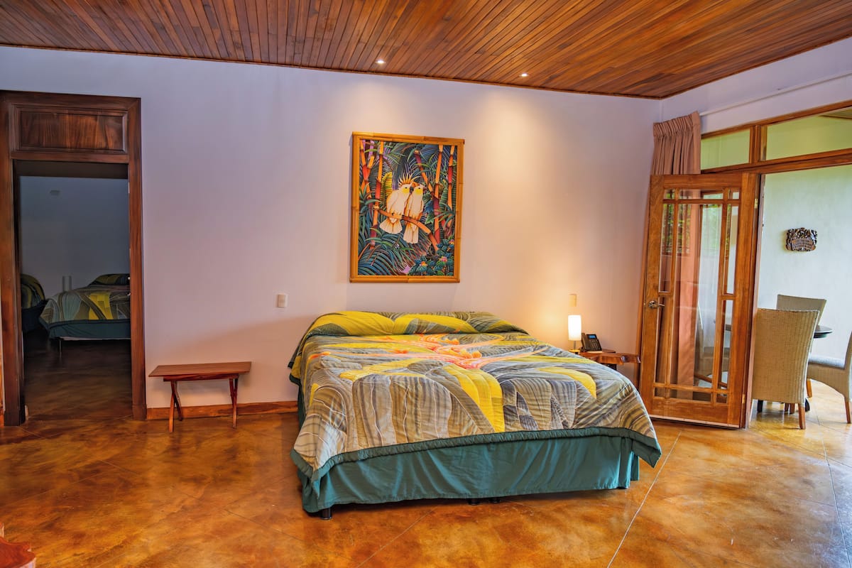 Une chambre avec un plafond en bois, un lit recouvert d’une courtepointe aux couleurs vives, un banc en bois et une porte donnant sur une autre pièce.