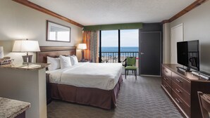 A hotel room with a large bed, a television, a desk with a lamp, a chair, and a view of the ocean. - Beach Quarters Resort (Virginia Beach)