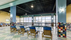 An indoor swimming pool area with wicker chairs, a tiled floor, and large windows offering a view of bicycles outside. - Beach Quarters Resort (Virginia Beach)