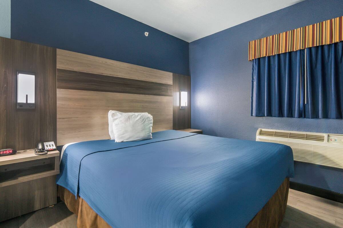 Habitación de hotel con una cama grande, cabecera de madera, mesita de noche con teléfono y despertador, y ventana con cortinas.