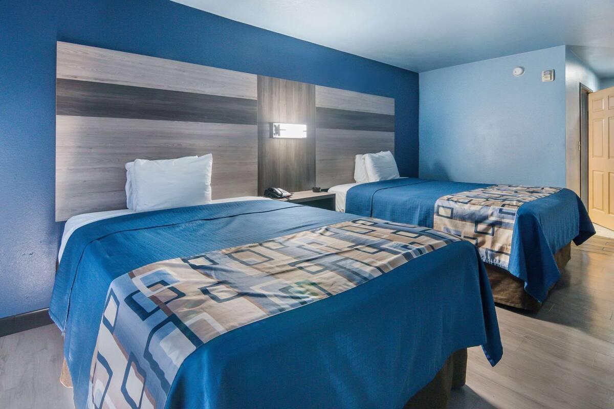 A hotel room with two beds, a wooden headboard, a bedside table with a lamp, and a door.