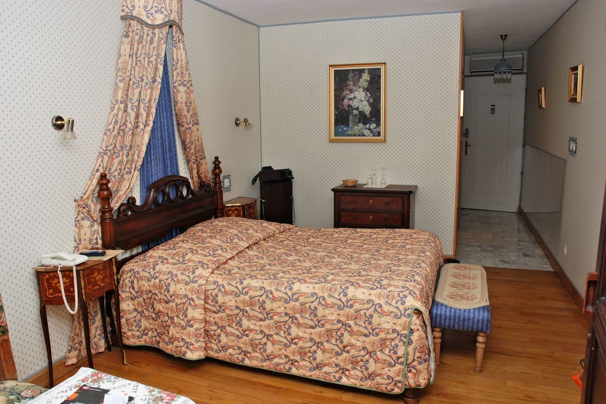 Ein Schlafzimmer mit einem hölzernen Bett, einem Nachttisch, einer Kommode, einer Bank, einem an der Wand befestigten Gemälde und einer Tür zu einem weiteren Raum.